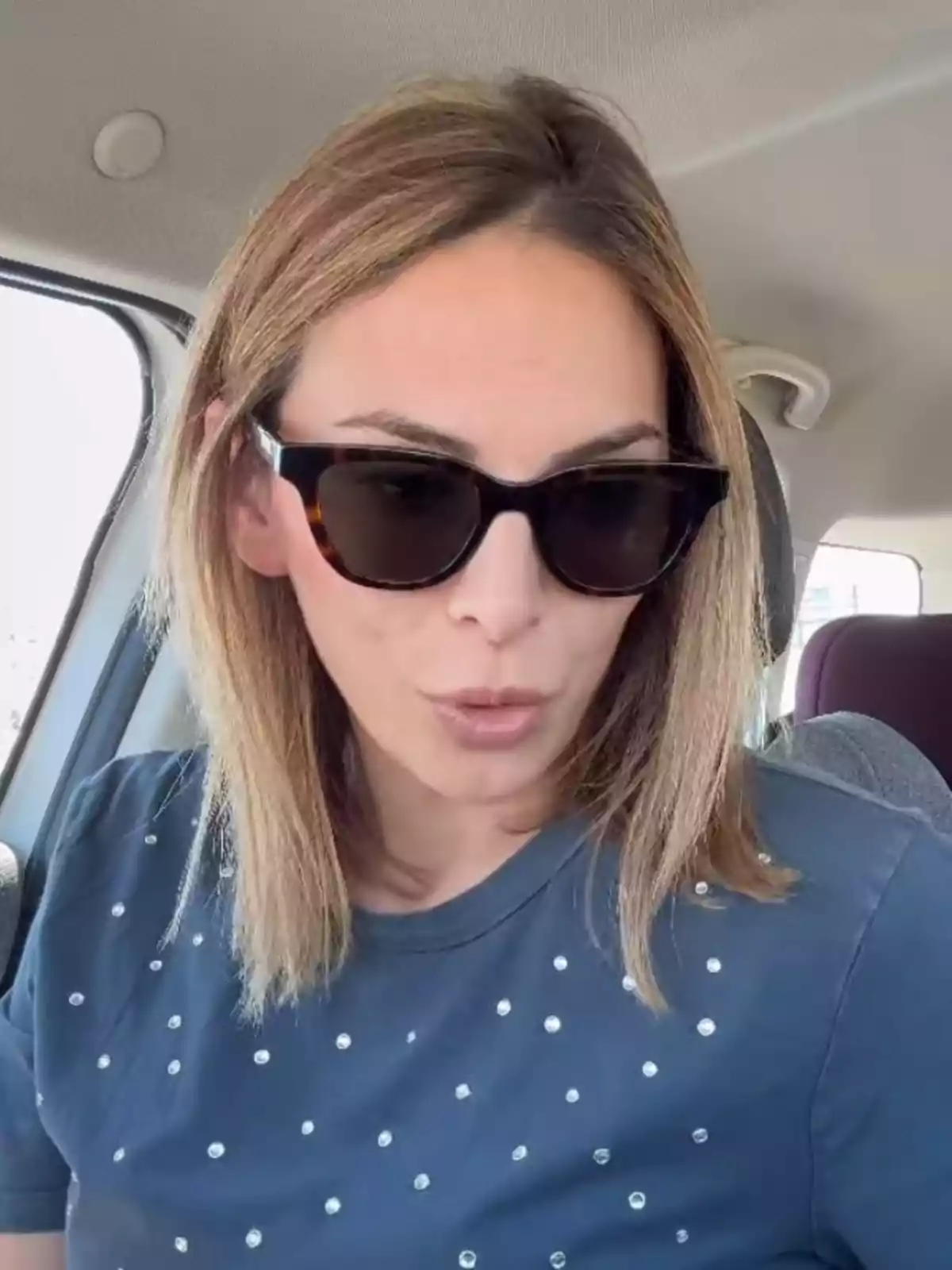 Irene Rosales con gafas de sol y cabello rubio está sentada en el interior de un automóvil, vistiendo una camiseta azul con detalles brillantes. Irene Rosales con gafas de sol y cabello rubio está sentada en el interior de un automóvil, vistiendo una camiseta azul con detalles brillantes.