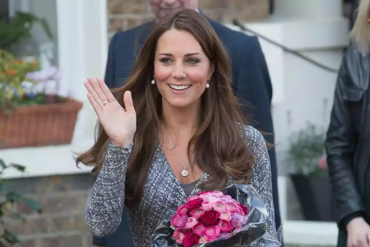 Una mujer sonriente con cabello largo y castaño saluda mientras sostiene un ramo de flores rosas. Una mujer sonriente con cabello largo y castaño saluda mientras sostiene un ramo de flores rosas.