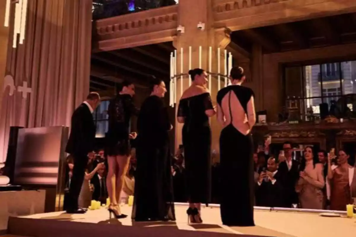Un grupo de personas elegantemente vestidas en un evento social en un salón con iluminación cálida y público observando. Un grupo de personas elegantemente vestidas en un evento social en un salón con iluminación cálida y público observando.
