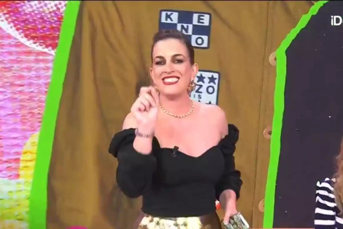 Leticia Requejo sonriente con un vestido negro de hombros descubiertos y falda dorada sostiene un objeto en la mano frente a un fondo colorido con letras y estrellas. Leticia Requejo sonriente con un vestido negro de hombros descubiertos y falda dorada sostiene un objeto en la mano frente a un fondo colorido con letras y estrellas.
