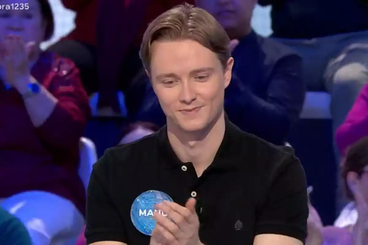 Manu joven con camisa negra aplaude mientras está sentado en un programa de televisión con personas en el fondo. Manu joven con camisa negra aplaude mientras está sentado en un programa de televisión con personas en el fondo.