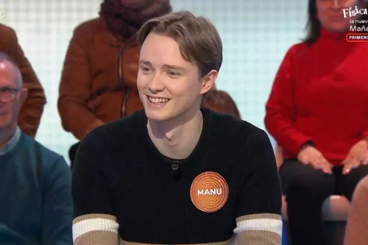 Manu sonriente con un suéter oscuro y una etiqueta que dice Manu sonriente con un suéter oscuro y una etiqueta que dice