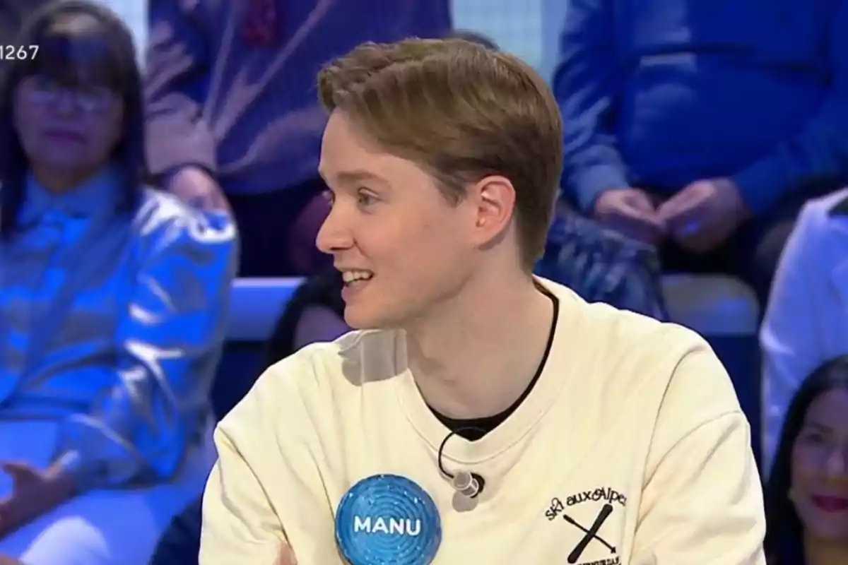 Manu joven con cabello claro y una camiseta blanca está sentado en un estudio de televisión con un micrófono en la camiseta y una placa que dice Manu joven con cabello claro y una camiseta blanca está sentado en un estudio de televisión con un micrófono en la camiseta y una placa que dice
