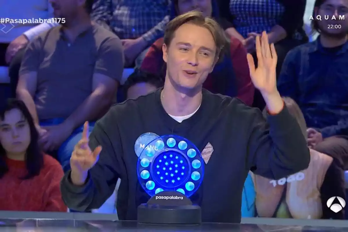 Manu en un programa de televisión levanta la mano mientras sonríe, con un público de fondo y un panel iluminado frente a él. Manu en un programa de televisión levanta la mano mientras sonríe, con un público de fondo y un panel iluminado frente a él.