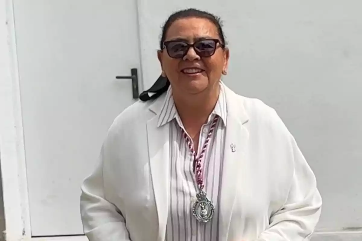 María del Monte con gafas de sol y una chaqueta blanca sonríe frente a una puerta blanca. María del Monte con gafas de sol y una chaqueta blanca sonríe frente a una puerta blanca.