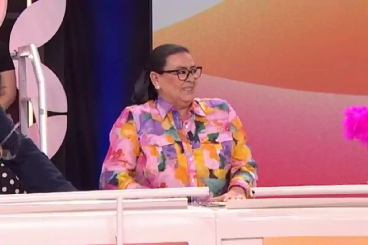 María del Monte con camisa colorida y gafas sonríe mientras está sentada en un set de televisión con fondo colorido. María del Monte con camisa colorida y gafas sonríe mientras está sentada en un set de televisión con fondo colorido.