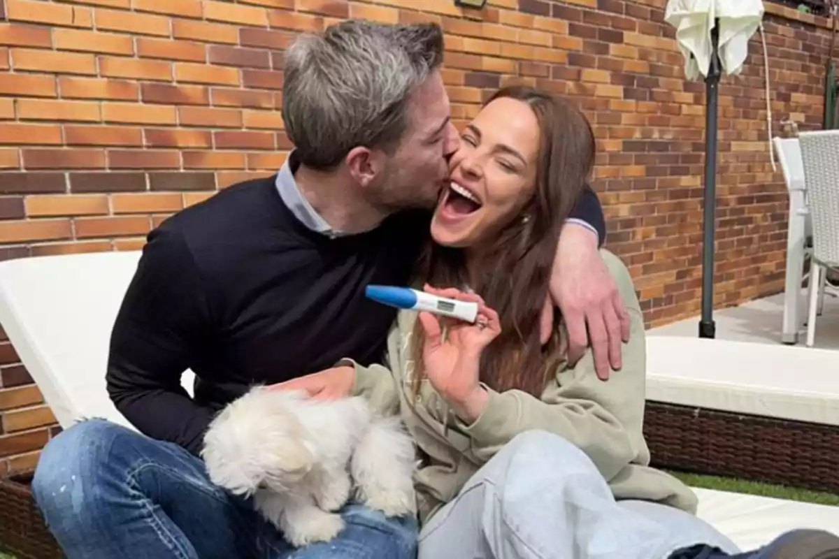 Marta Peñate y Tony Spina sonrientes se abraza mientras la mujer sostiene una prueba de embarazo y un pequeño perro blanco está en su regazo.