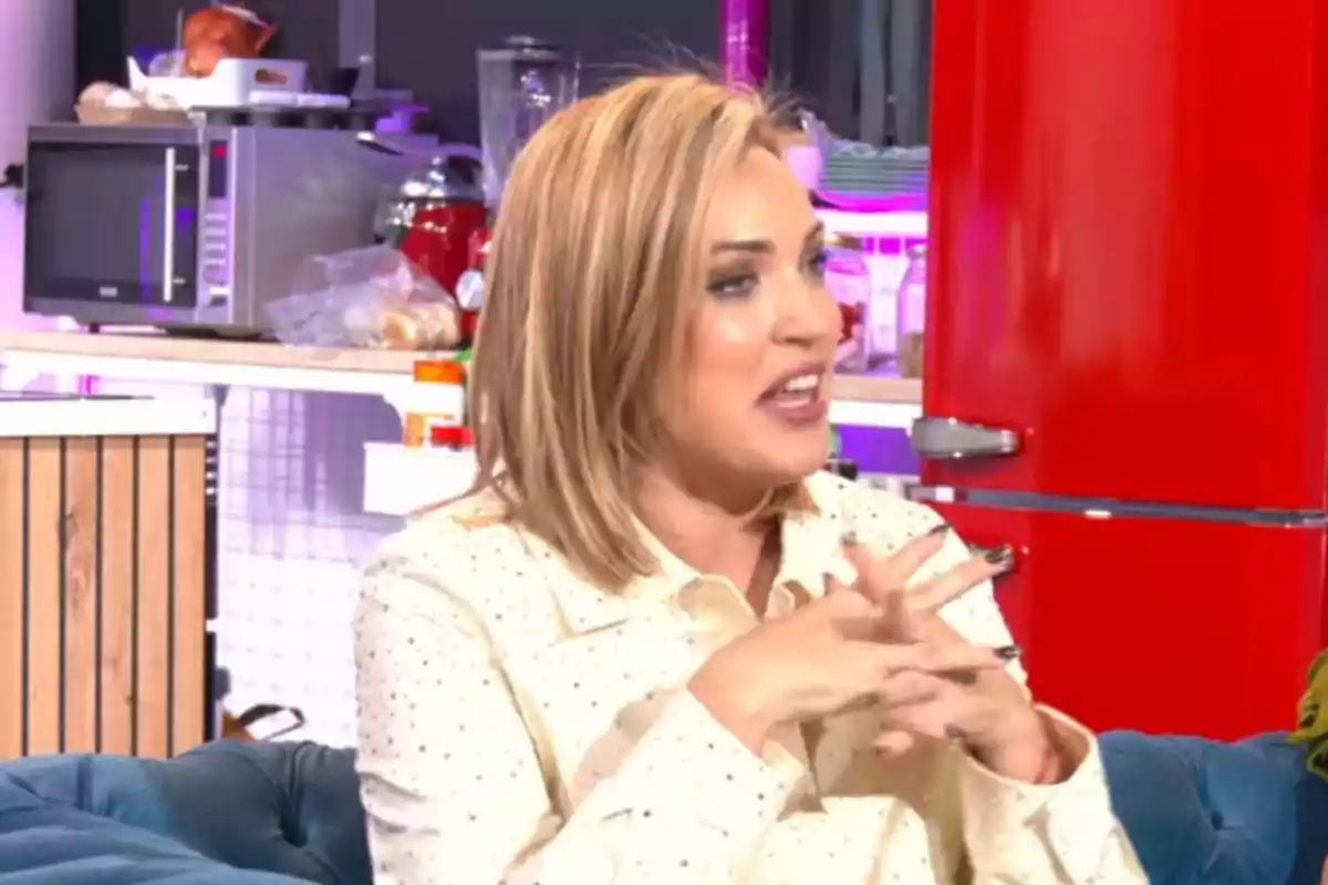 Marta Riesco con cabello rubio y camisa clara está sentada en un sofá azul en un ambiente de cocina moderna con electrodomésticos y un refrigerador rojo al fondo. Marta Riesco con cabello rubio y camisa clara está sentada en un sofá azul en un ambiente de cocina moderna con electrodomésticos y un refrigerador rojo al fondo.
