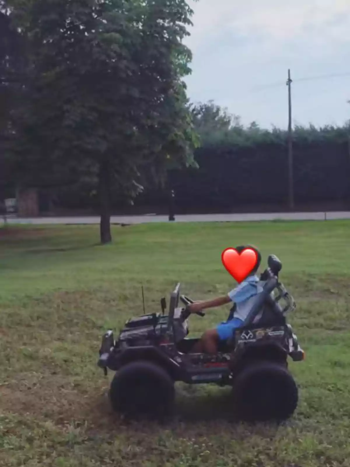 Niño conduciendo un coche de juguete negro en un campo de césped con árboles al fondo y un emoji de corazón cubriendo su rostro Niño conduciendo un coche de juguete negro en un campo de césped con árboles al fondo y un emoji de corazón cubriendo su rostro