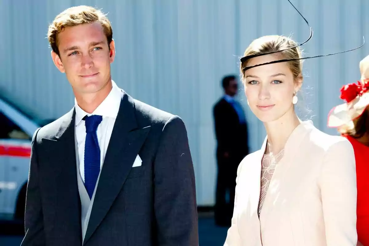 Una pareja elegantemente vestida en un evento al aire libre, con el hombre usando un traje oscuro y corbata azul, y la mujer con un sombrero decorativo y un atuendo claro. Una pareja elegantemente vestida en un evento al aire libre, con el hombre usando un traje oscuro y corbata azul, y la mujer con un sombrero decorativo y un atuendo claro.