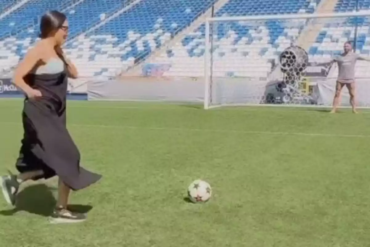 Pilar Rubio con vestido negro y zapatillas se prepara para patear un balón de fútbol en un estadio mientras Sergio Ramos está en la portería. Pilar Rubio con vestido negro y zapatillas se prepara para patear un balón de fútbol en un estadio mientras Sergio Ramos está en la portería.