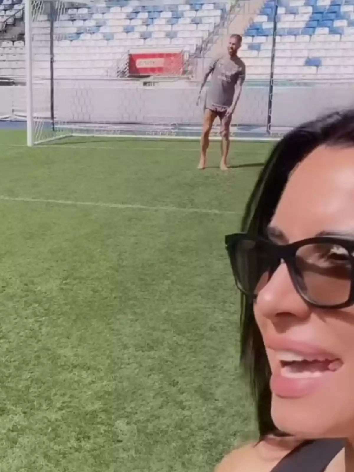 Pilar Rubio con gafas de sol sonríe en primer plano mientras Sergio Ramos está descalzo en una portería de fútbol en el fondo. Pilar Rubio con gafas de sol sonríe en primer plano mientras Sergio Ramos está descalzo en una portería de fútbol en el fondo.
