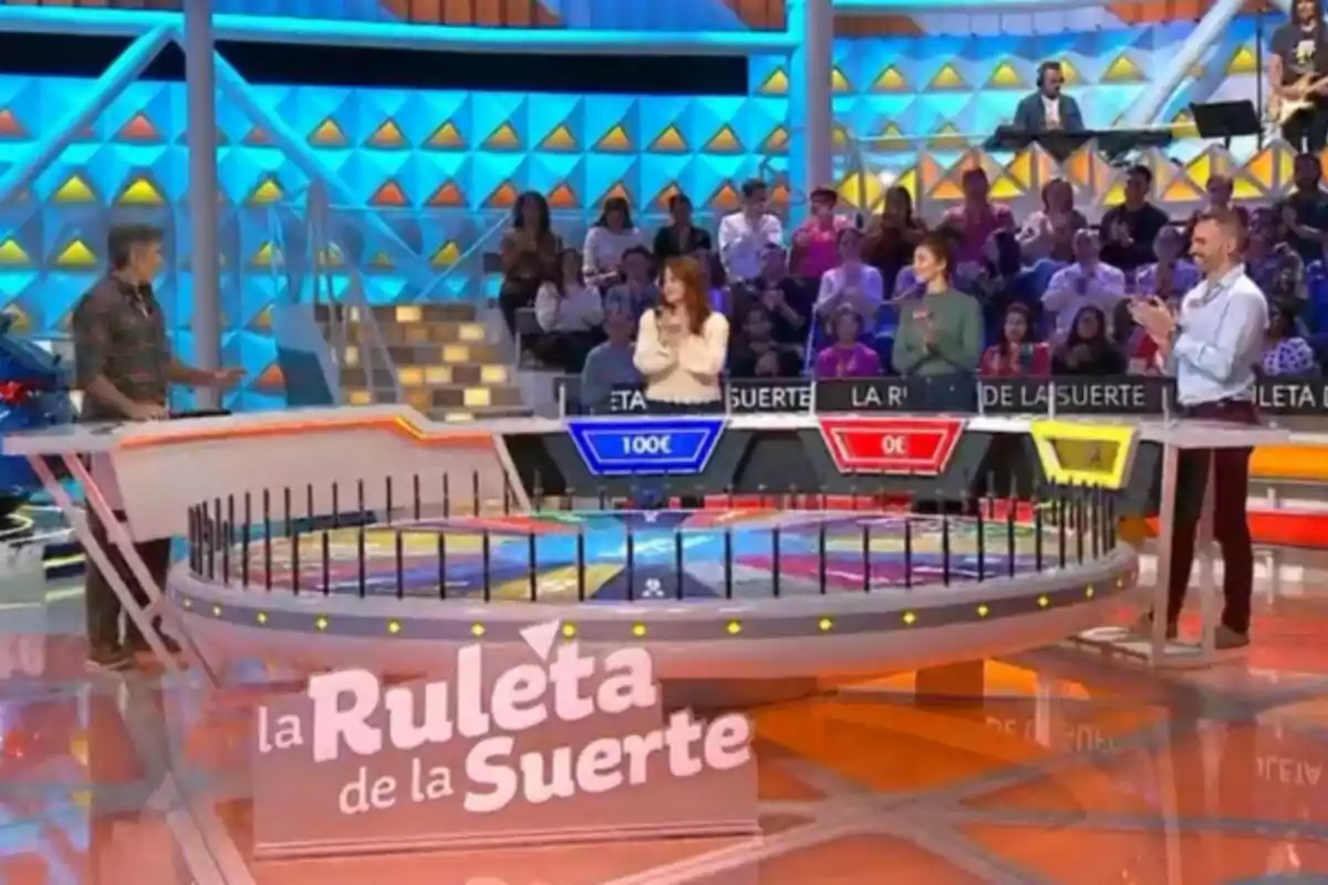 Plano general del plató de un programa de televisión, que es 'La Ruleta de la Suerte' con Jorge Fernández y tres concursantes de pie alrededor de una gran ruleta en un estudio colorido con público en el fondo. Plano general del plató de un programa de televisión, que es 'La Ruleta de la Suerte' con Jorge Fernández y tres concursantes de pie alrededor de una gran ruleta en un estudio colorido con público en el fondo.