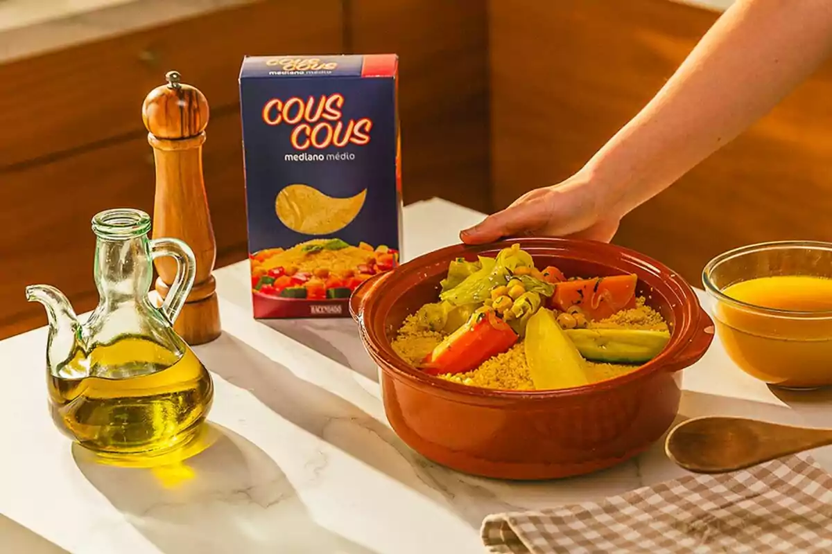 Un plato de cuscús con verduras en un recipiente de barro junto a una caja de cuscús, una botella de aceite de oliva, un molinillo de pimienta y un tazón de caldo sobre una mesa. Un plato de cuscús con verduras en un recipiente de barro junto a una caja de cuscús, una botella de aceite de oliva, un molinillo de pimienta y un tazón de caldo sobre una mesa.