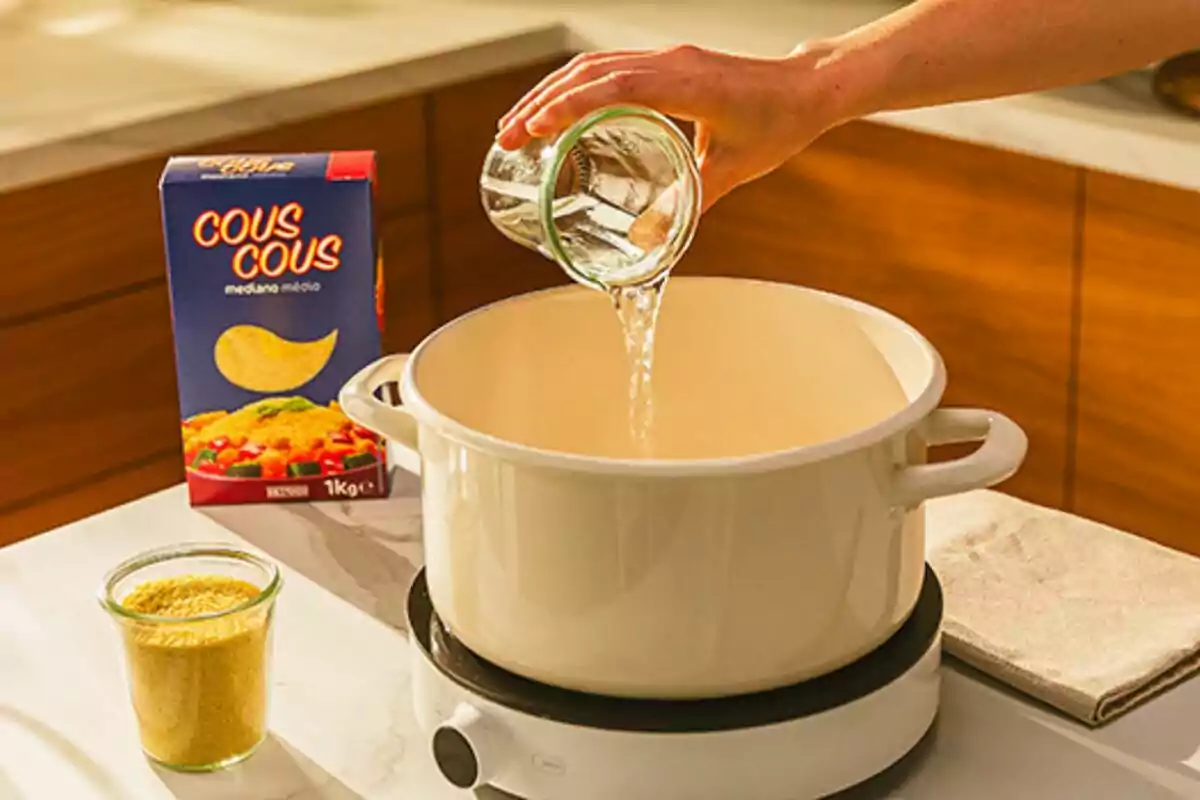 Una mano vertiendo agua en una olla blanca sobre una estufa, con una caja de cuscús y un frasco de cuscús en la mesa. Una mano vertiendo agua en una olla blanca sobre una estufa, con una caja de cuscús y un frasco de cuscús en la mesa.