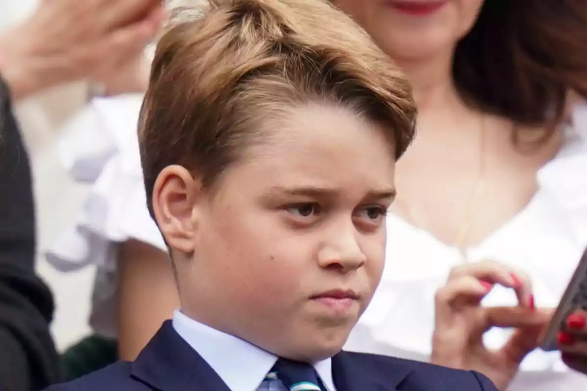 Un niño con expresión seria, vestido con traje y corbata, rodeado de personas en un evento. Un niño con expresión seria, vestido con traje y corbata, rodeado de personas en un evento.