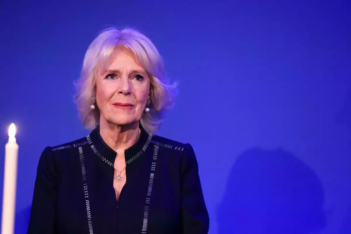 Mujer de cabello rubio y corto con expresión seria vestida de negro frente a un fondo azul y una vela encendida a su lado Mujer de cabello rubio y corto con expresión seria vestida de negro frente a un fondo azul y una vela encendida a su lado