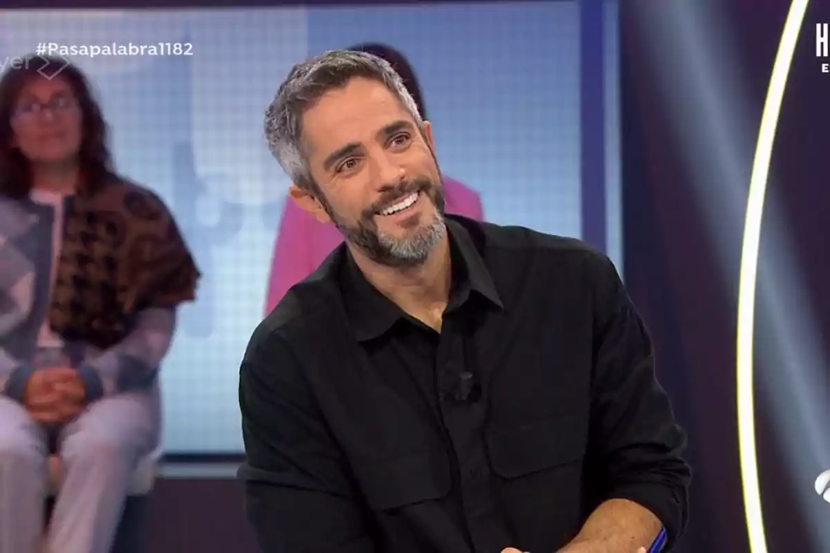 Roberto Leal sonriente con barba y camisa negra en un programa de televisión. Roberto Leal sonriente con barba y camisa negra en un programa de televisión.