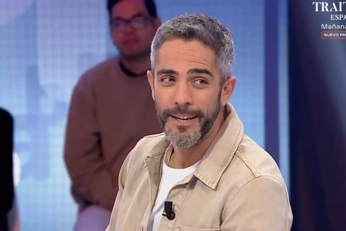 Roberto Leal con barba y cabello canoso está sonriendo mientras participa en un programa de televisión con un fondo azul y otra persona desenfocada detrás. Roberto Leal con barba y cabello canoso está sonriendo mientras participa en un programa de televisión con un fondo azul y otra persona desenfocada detrás.