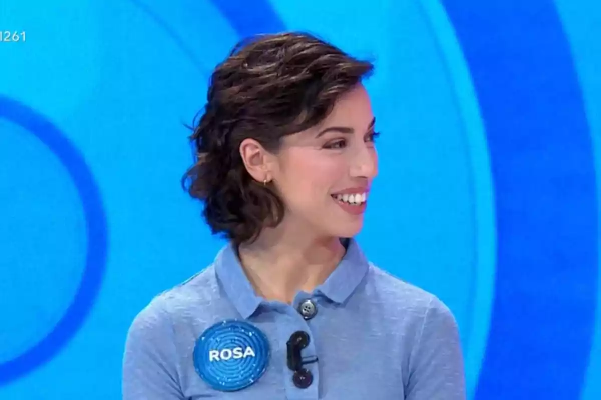 Rosa con cabello corto y oscuro sonríe mientras lleva una camisa azul con un botón y un micrófono en un fondo azul. Rosa con cabello corto y oscuro sonríe mientras lleva una camisa azul con un botón y un micrófono en un fondo azul.