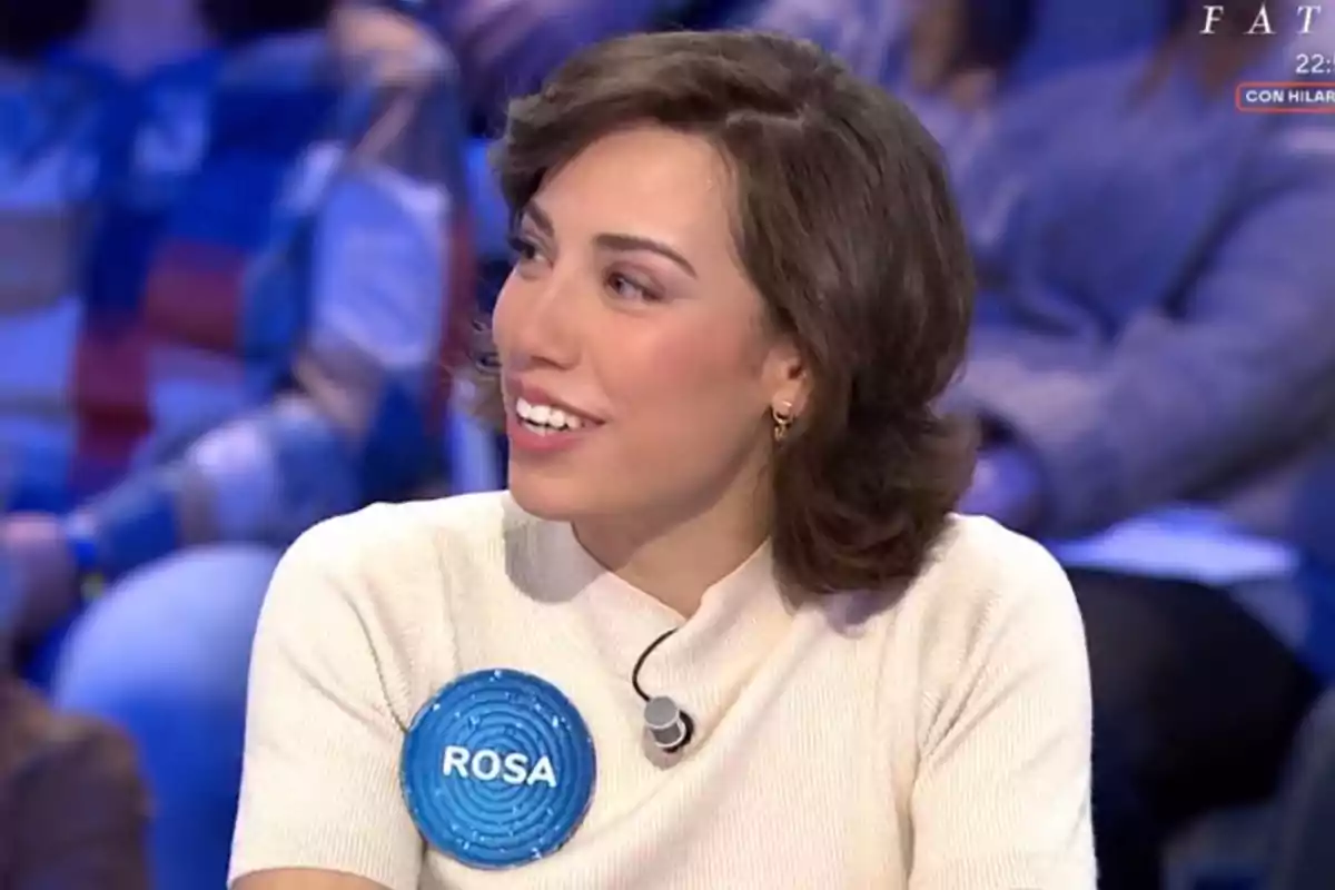 Rosa con cabello corto y oscuro, sonriendo y mirando hacia un lado, lleva un micrófono en la ropa y un distintivo azul con el nombre Rosa con cabello corto y oscuro, sonriendo y mirando hacia un lado, lleva un micrófono en la ropa y un distintivo azul con el nombre