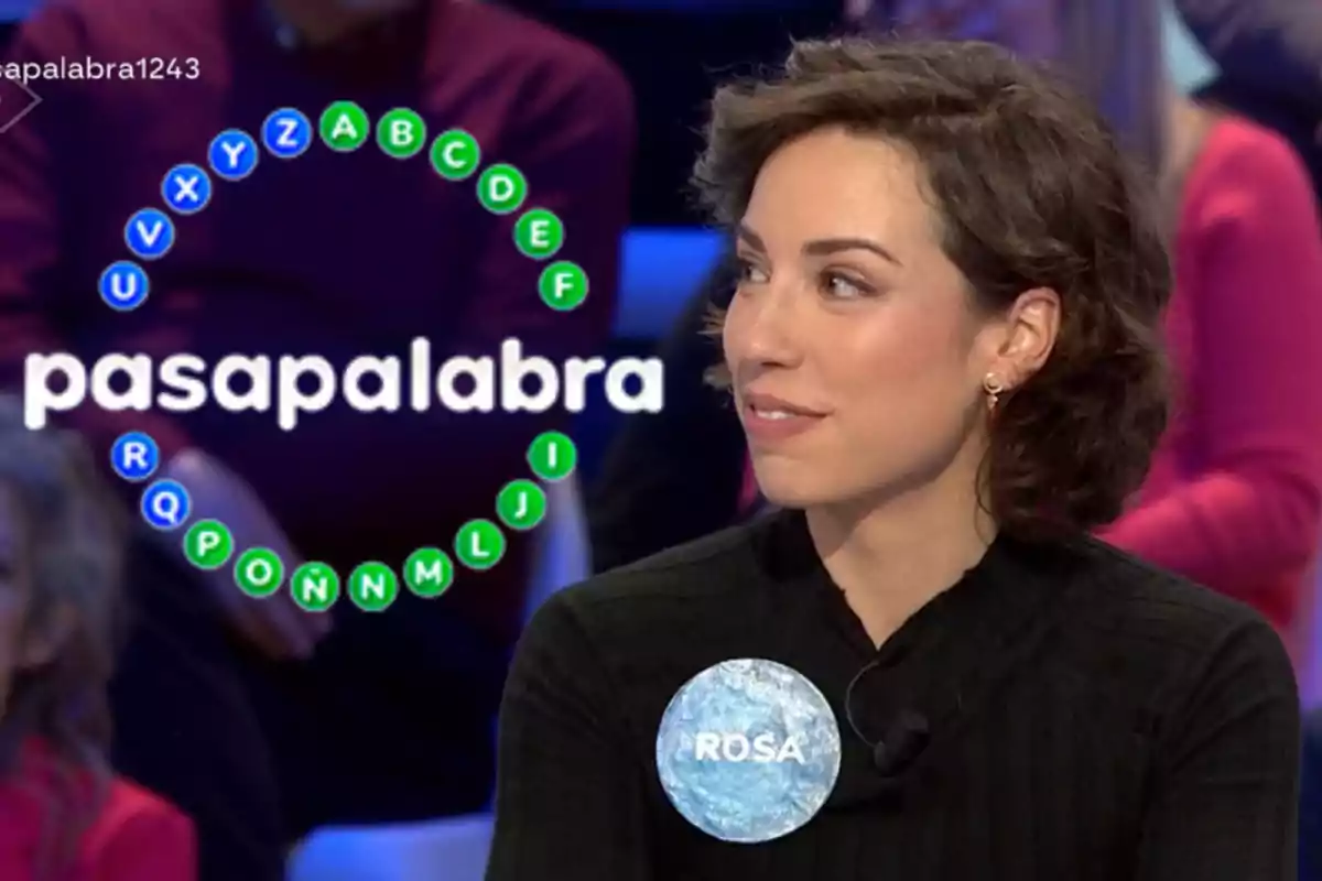 Rosa en un programa de televisión con un círculo de letras en el fondo y un micrófono en la ropa.