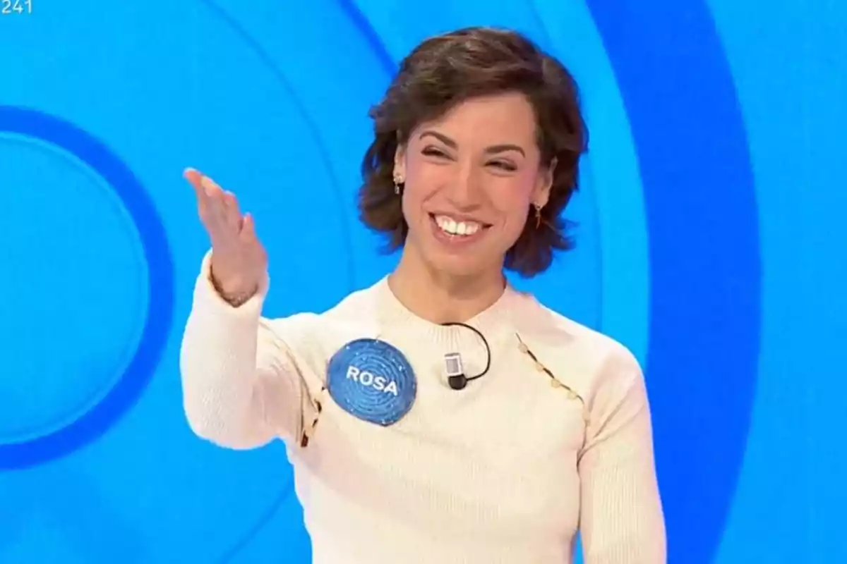 Rosa sonriente con cabello corto y oscuro, vestida con un suéter claro, está en un set de televisión con un fondo azul. Rosa sonriente con cabello corto y oscuro, vestida con un suéter claro, está en un set de televisión con un fondo azul.
