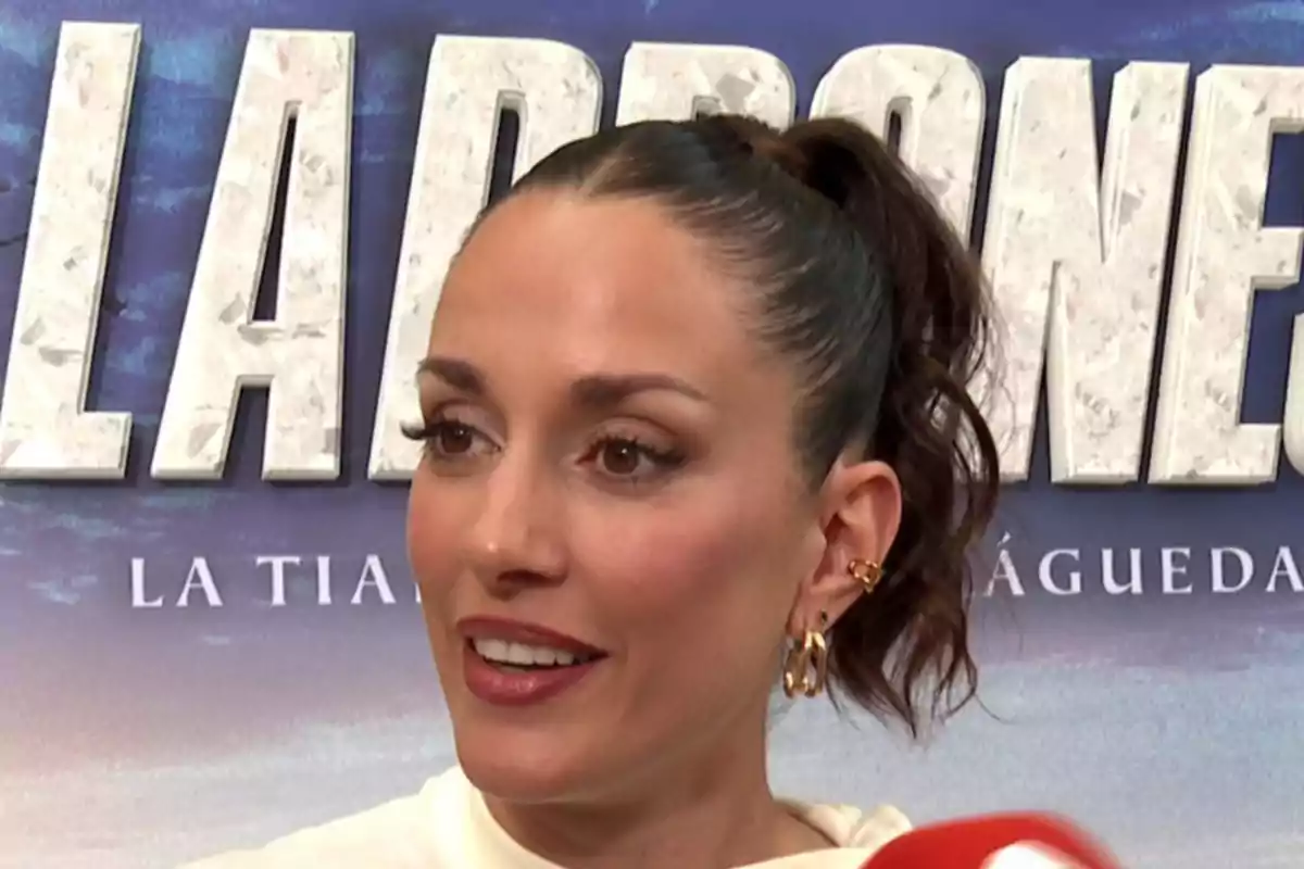 Silvia Alonso de cabello recogido con coleta alta y pendientes dorados sonriendo frente a un fondo con letras grandes blancas Silvia Alonso de cabello recogido con coleta alta y pendientes dorados sonriendo frente a un fondo con letras grandes blancas
