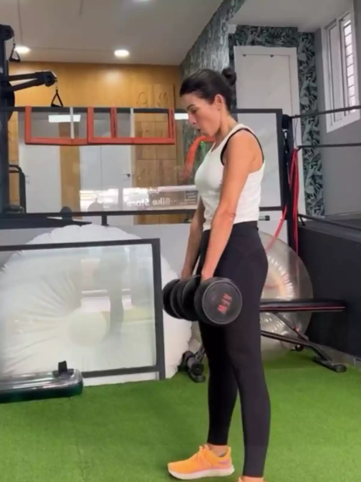 Virginia Troconis levantando pesas en un gimnasio con piso de césped artificial y equipo de ejercicio en el fondo. Virginia Troconis levantando pesas en un gimnasio con piso de césped artificial y equipo de ejercicio en el fondo.