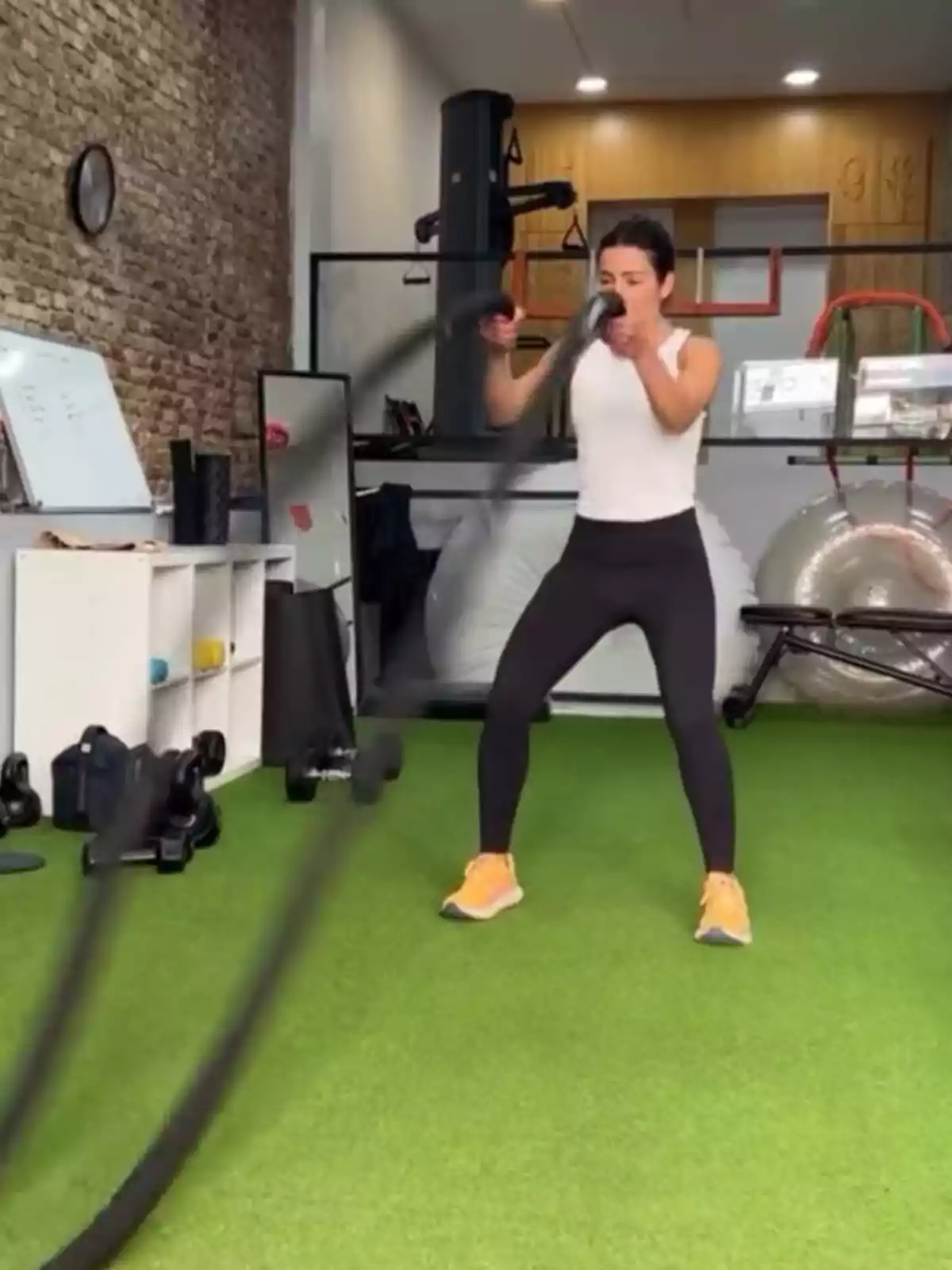 Virginia Troconis haciendo ejercicio con cuerdas de batalla en un gimnasio con piso de césped artificial y equipo de entrenamiento alrededor. Virginia Troconis haciendo ejercicio con cuerdas de batalla en un gimnasio con piso de césped artificial y equipo de entrenamiento alrededor.