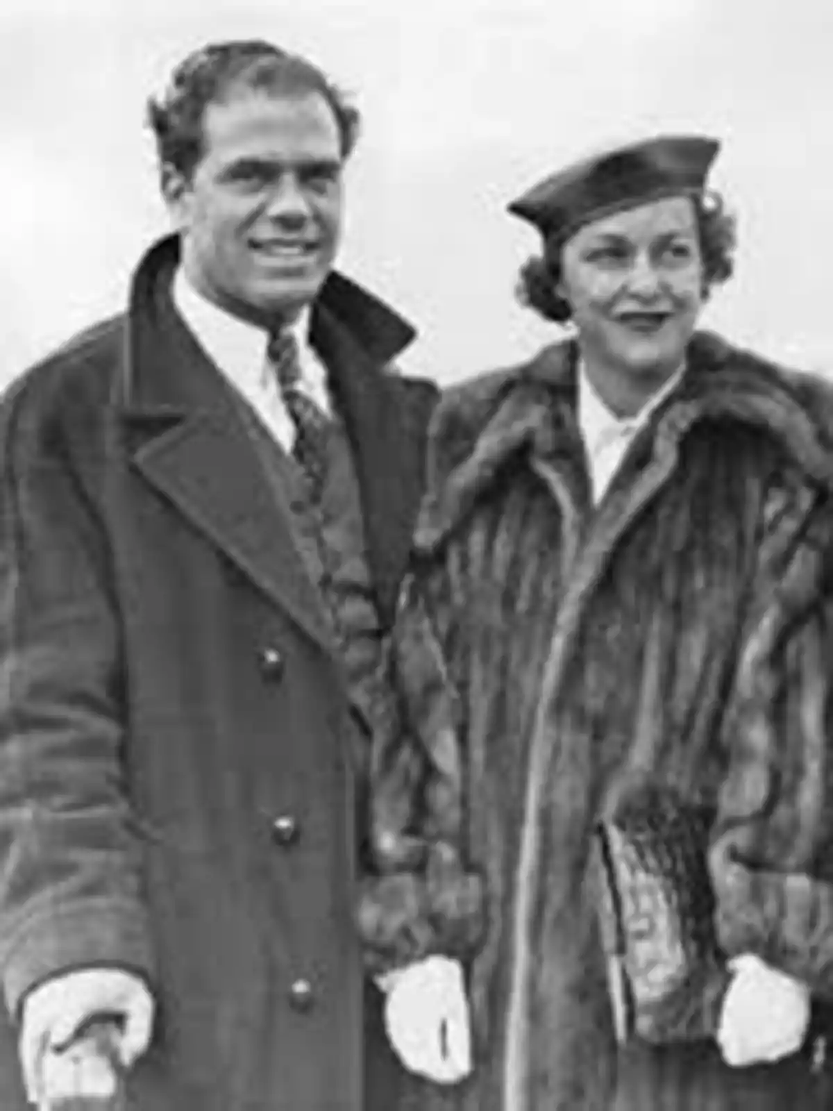 Una pareja de pie, ambos con abrigos elegantes, el hombre lleva un abrigo oscuro y la mujer un abrigo de piel, ambos sonríen mientras miran hacia adelante. Una pareja de pie, ambos con abrigos elegantes, el hombre lleva un abrigo oscuro y la mujer un abrigo de piel, ambos sonríen mientras miran hacia adelante.