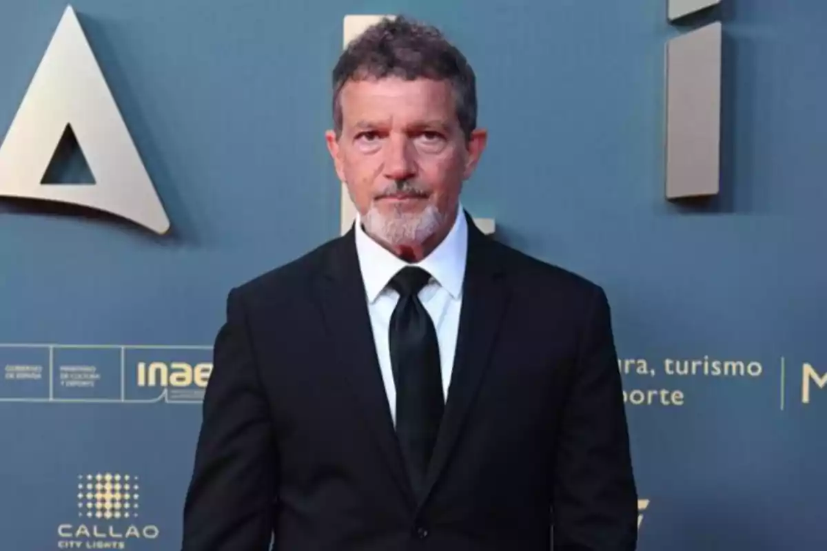 Un hombre con traje negro y corbata posa frente a un fondo azul con letras y logotipos.