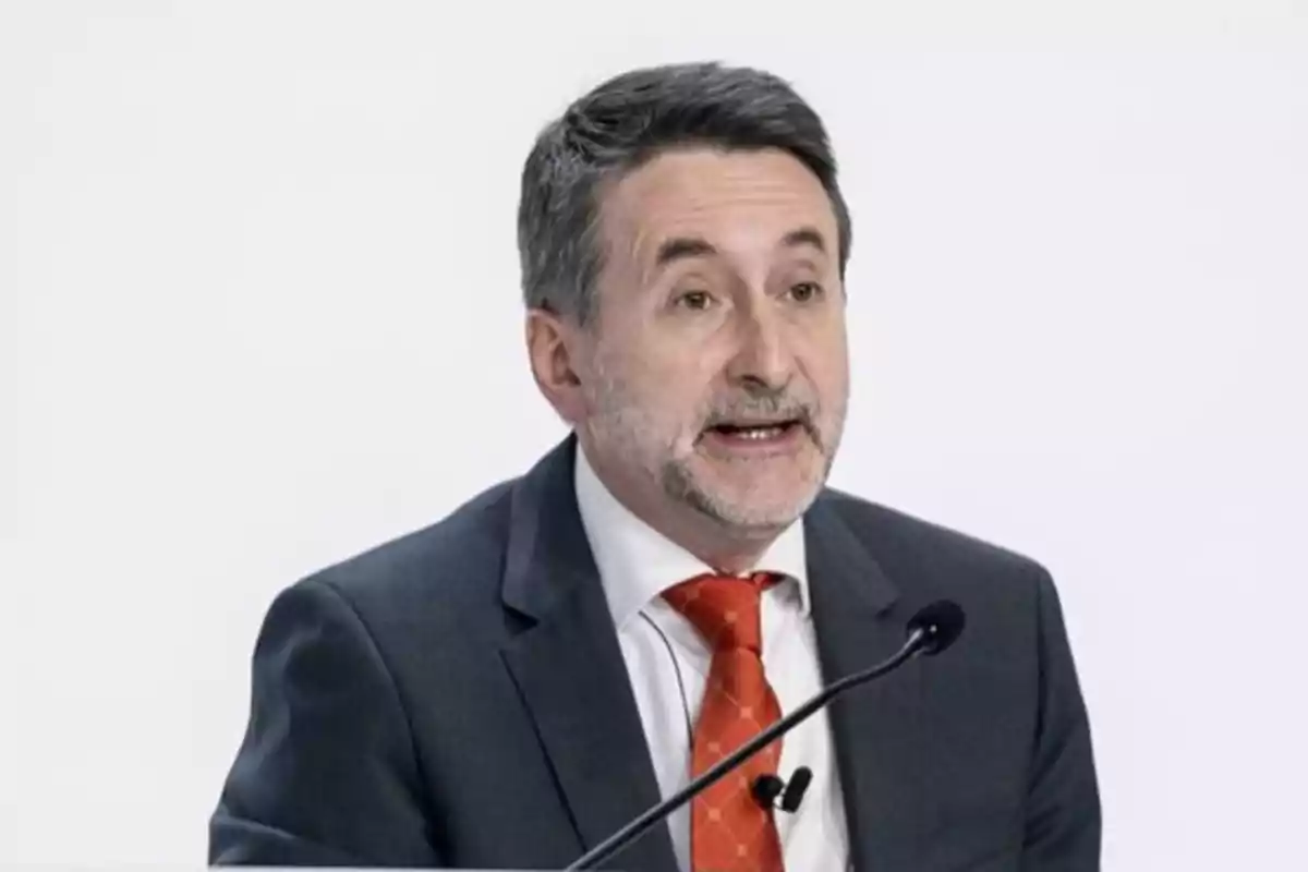 Hombre de mediana edad con barba y cabello canoso hablando en un micrófono mientras viste un traje gris y corbata roja. Hombre de mediana edad con barba y cabello canoso hablando en un micrófono mientras viste un traje gris y corbata roja.