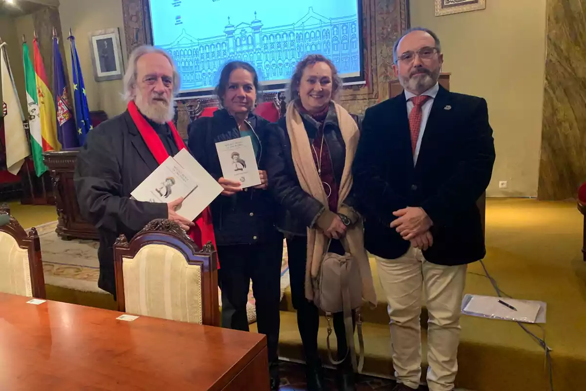 Cuatro personas posan juntas en una sala con banderas y un proyector al fondo, dos de ellas sostienen libros.