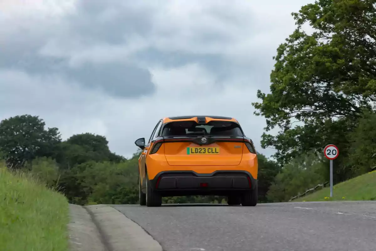 Un MG 4 EV naranja circula por una carretera rodeada de árboles y un cielo nublado.