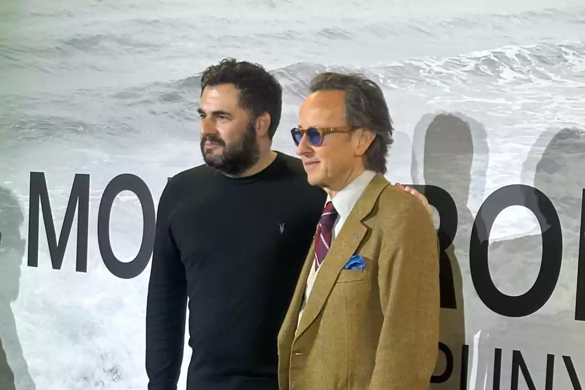 Dos hombres posan juntos frente a un fondo con letras grandes y un diseño de olas.