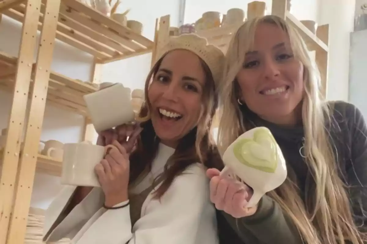 Dos personas sonrientes sosteniendo tazas de cerámica en un taller con estantes llenos de más tazas.