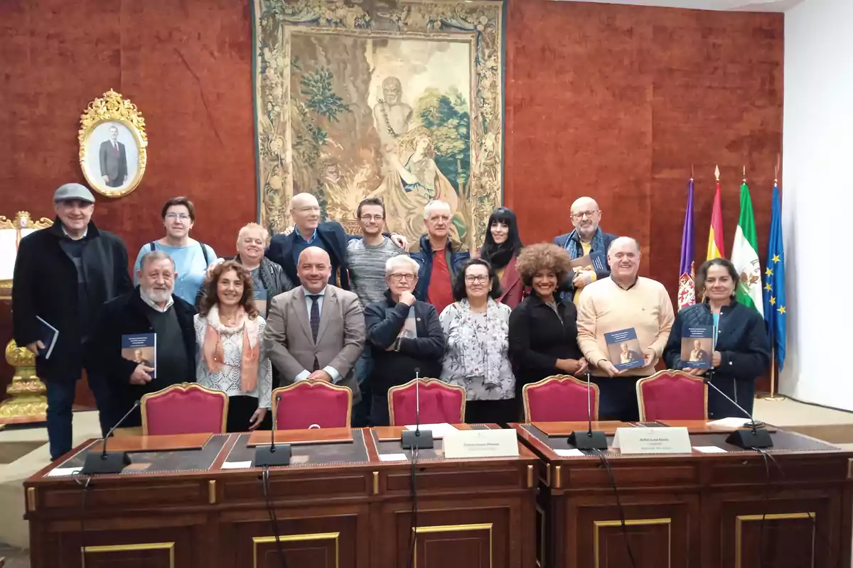 Un grupo de personas posa en una sala con decoración elegante, incluyendo un tapiz y varias banderas, mientras sostienen libros.