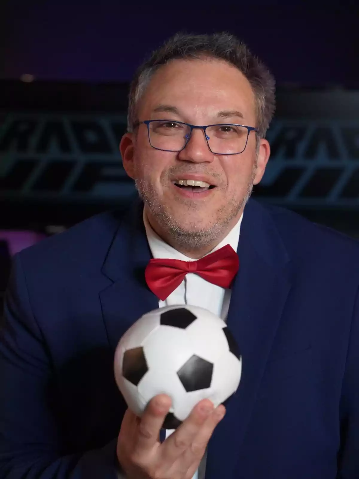 Un hombre con gafas y pajarita roja sostiene un balón de fútbol pequeño mientras sonríe a la cámara.