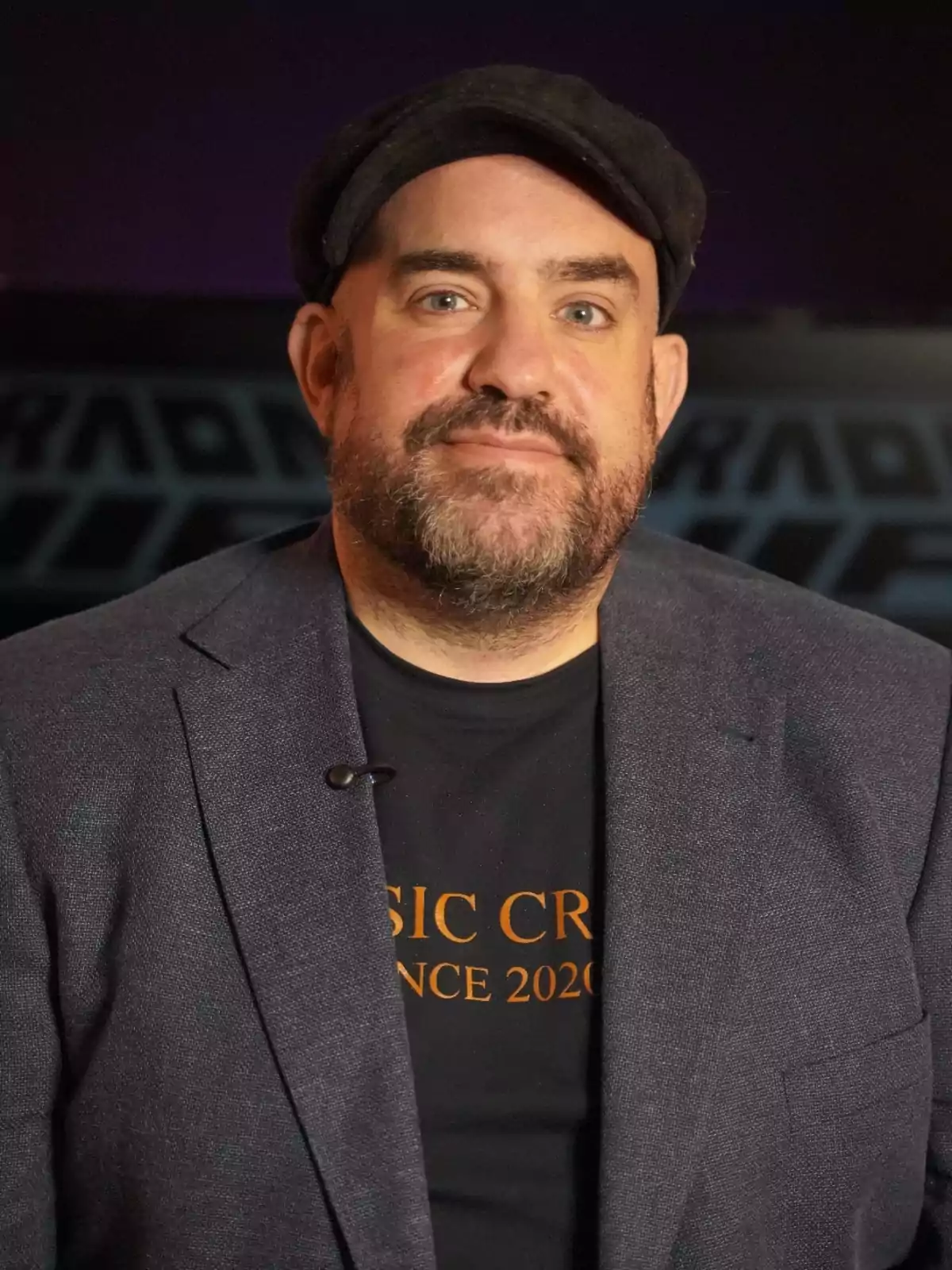 Un hombre con barba y gorra negra lleva una camiseta negra y una chaqueta azul oscuro.