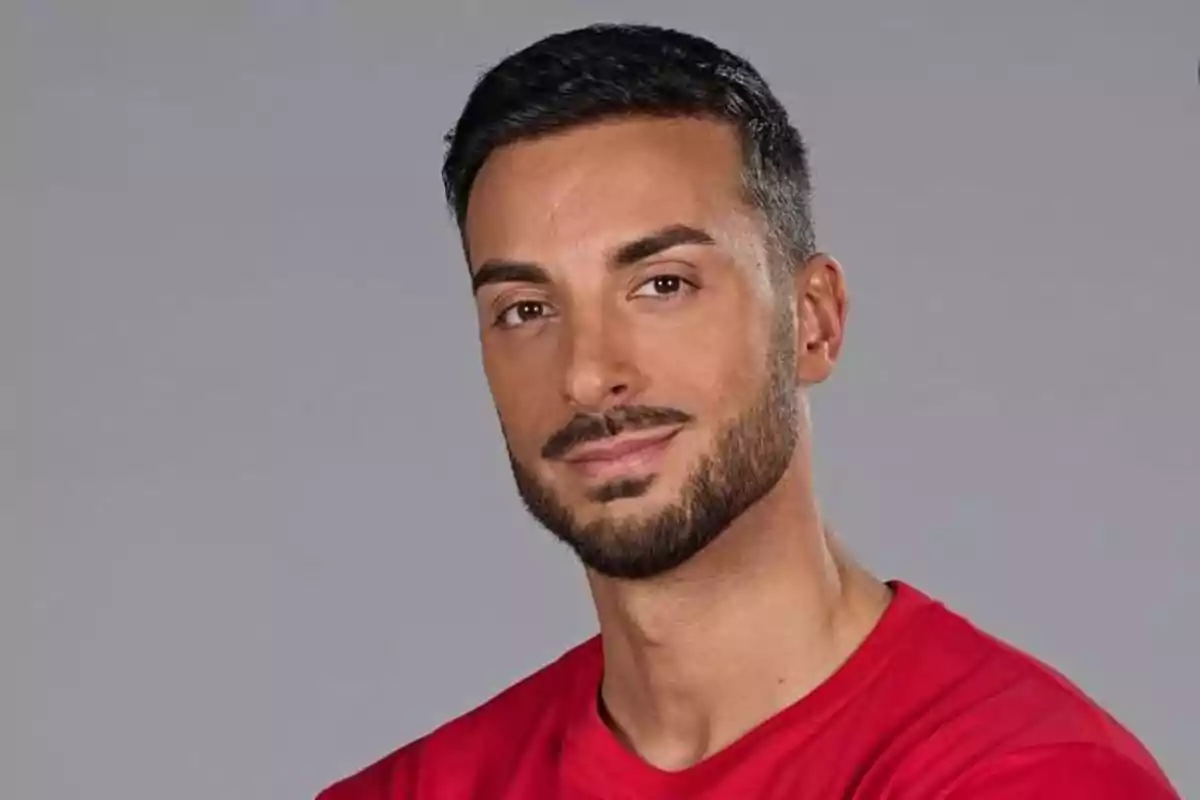 Hombre joven con barba y cabello corto oscuro vistiendo una camiseta roja sobre fondo gris Hombre joven con barba y cabello corto oscuro vistiendo una camiseta roja sobre fondo gris