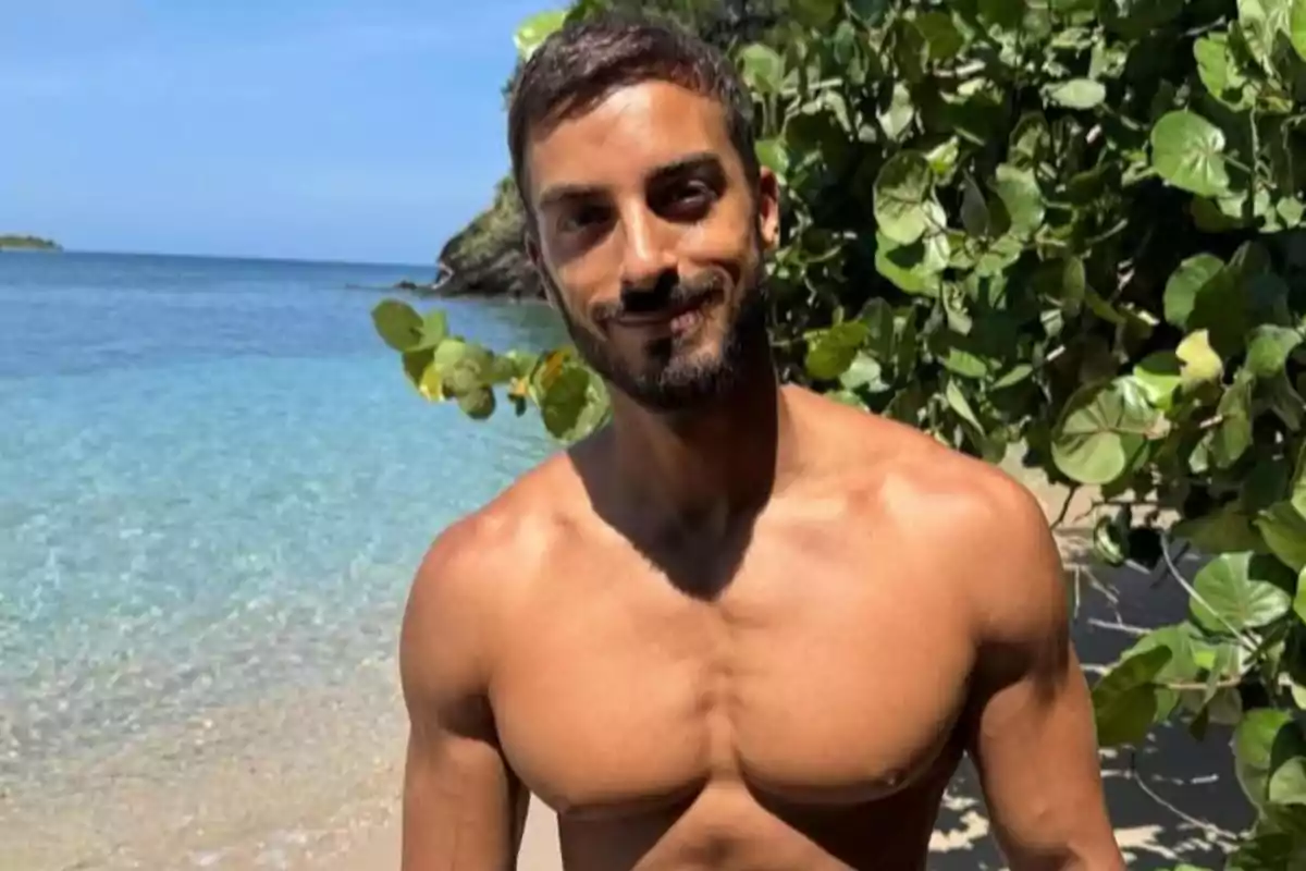 Hombre joven sin camiseta sonriendo en la playa con agua cristalina y vegetación al fondo