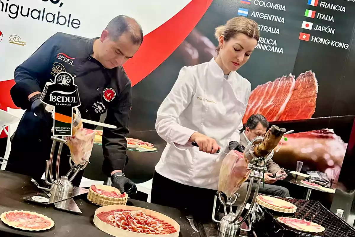 Dos personas en un evento gastronómico cortan jamón serrano en un stand decorado con imágenes de jamón y banderas de varios países.