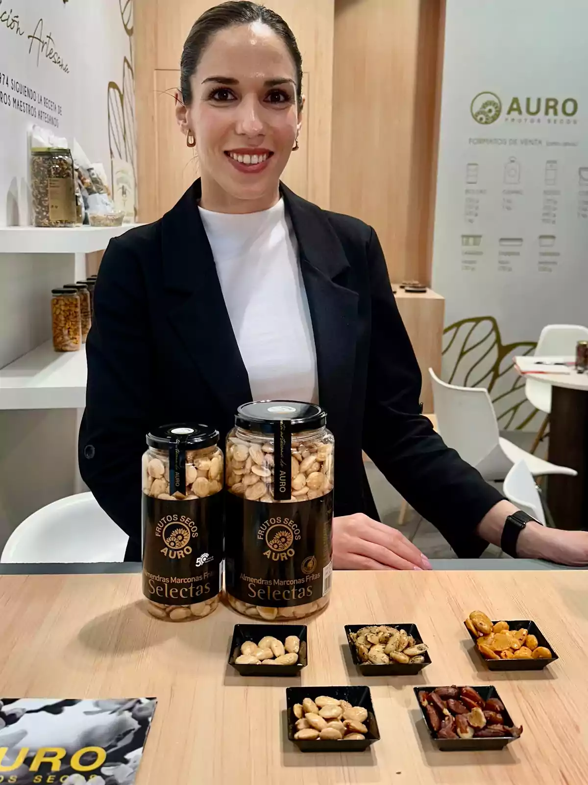 Una mujer sonriente está de pie detrás de una mesa que exhibe frascos de almendras y otros frutos secos de la marca Auro en un entorno de feria o exposición. Una mujer sonriente está de pie detrás de una mesa que exhibe frascos de almendras y otros frutos secos de la marca Auro en un entorno de feria o exposición.