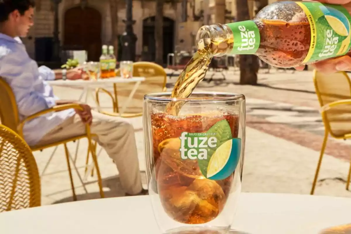 Una persona vierte té helado Fuze Tea en un vaso transparente en una terraza al aire libre.