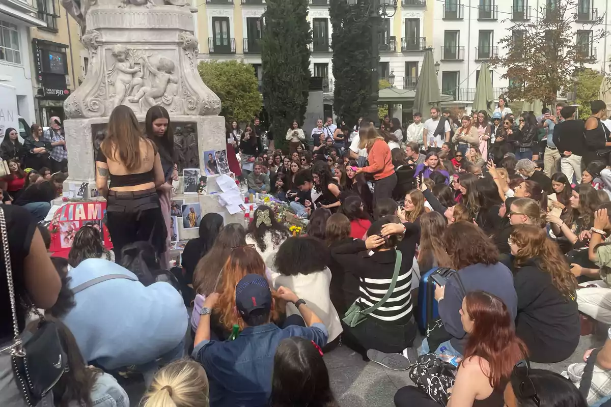 Un grupo de personas se reúne alrededor de un monumento en una plaza, donde han colocado fotos, flores y mensajes en un ambiente de conmemoración.