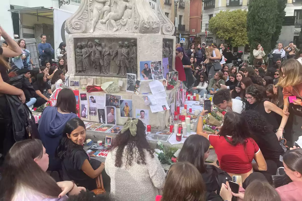 Un grupo de personas se reúne alrededor de un monumento decorado con fotos, velas y flores en un homenaje en un espacio público.