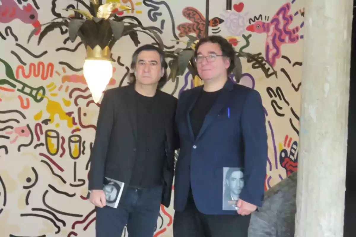 Dos hombres posan juntos frente a una pared con un diseño artístico colorido, ambos sosteniendo libros en sus manos.