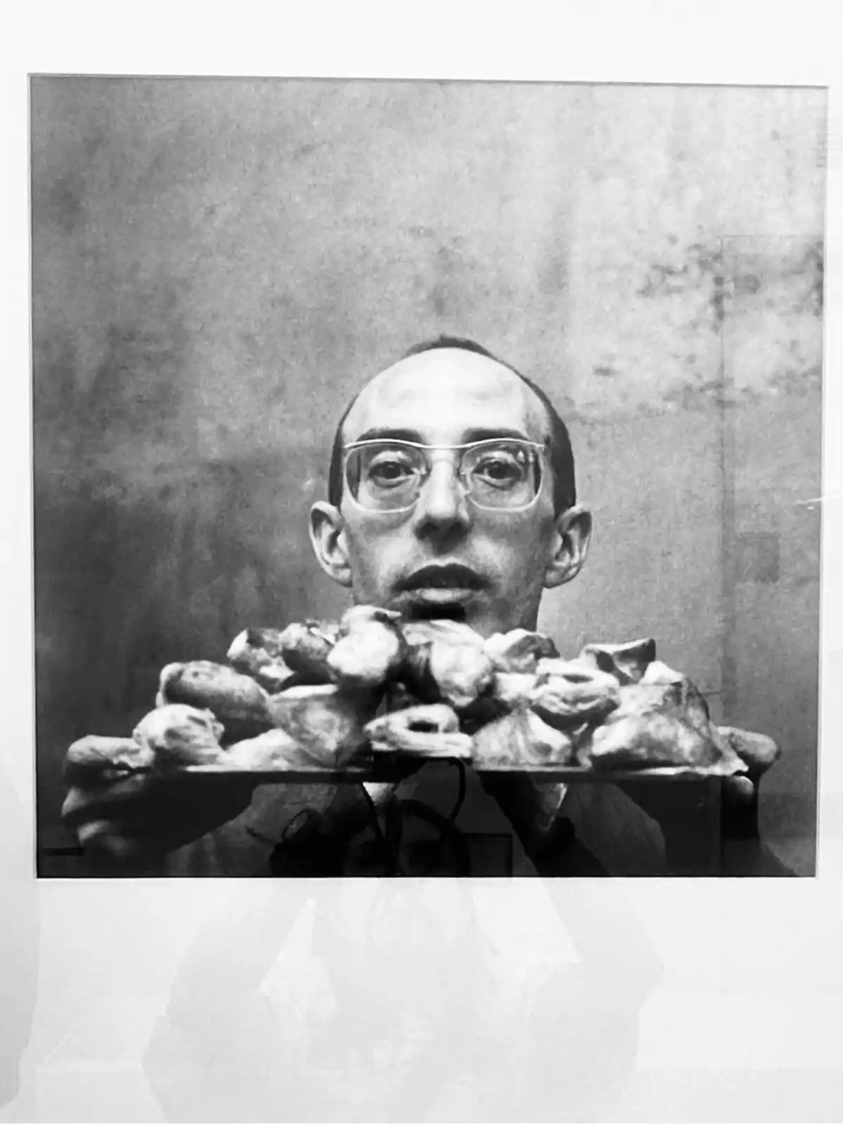 Hombre con gafas sostiene una bandeja llena de piezas de pan o bollos y mira fijamente hacia la cámara en una fotografía en blanco y negro Hombre con gafas sostiene una bandeja llena de piezas de pan o bollos y mira fijamente hacia la cámara en una fotografía en blanco y negro
