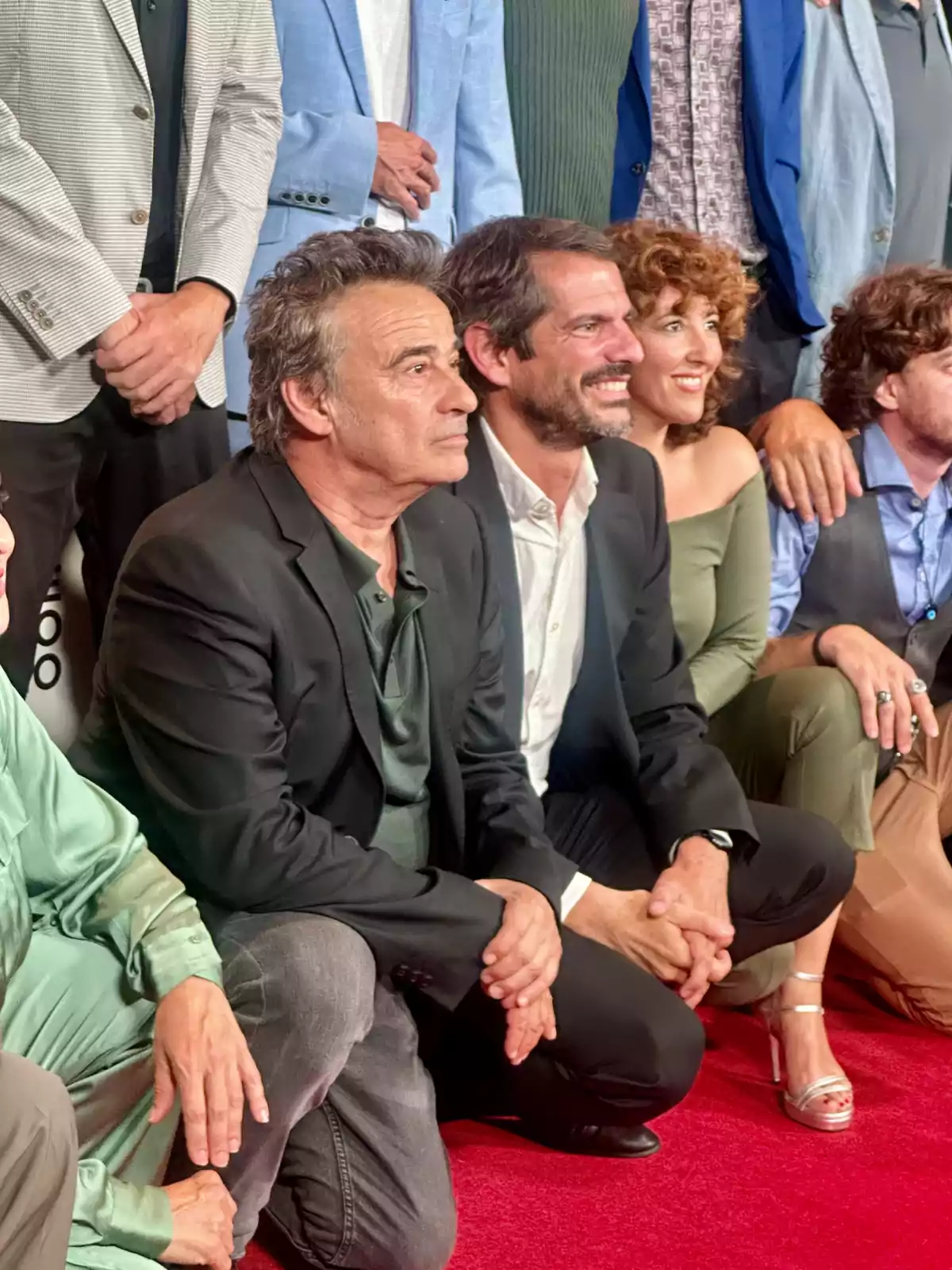 Un grupo de personas posando juntas en una alfombra roja, algunas de ellas están arrodilladas y otras de pie, todos vestidos de manera formal.